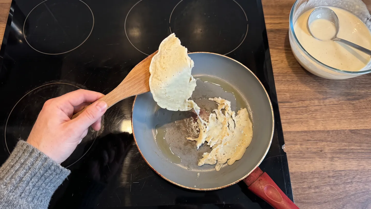Aus der Sicht der Köchin: Ein Pfannkuchen reißt in einer alten Pfanne, eine Hälfte liegt auf dem Holzwender, die andere klebt am Boden (Symbolbild, mit KI erstellt)